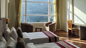 Quarto de hotel com duas camas, uma escrivaninha, uma cadeira, um ventilador de teto e uma grande janela com vista.