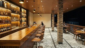 Uma área de bar com um longo balcão de madeira, várias prateleiras com garrafas de bebidas e banquetas de couro.