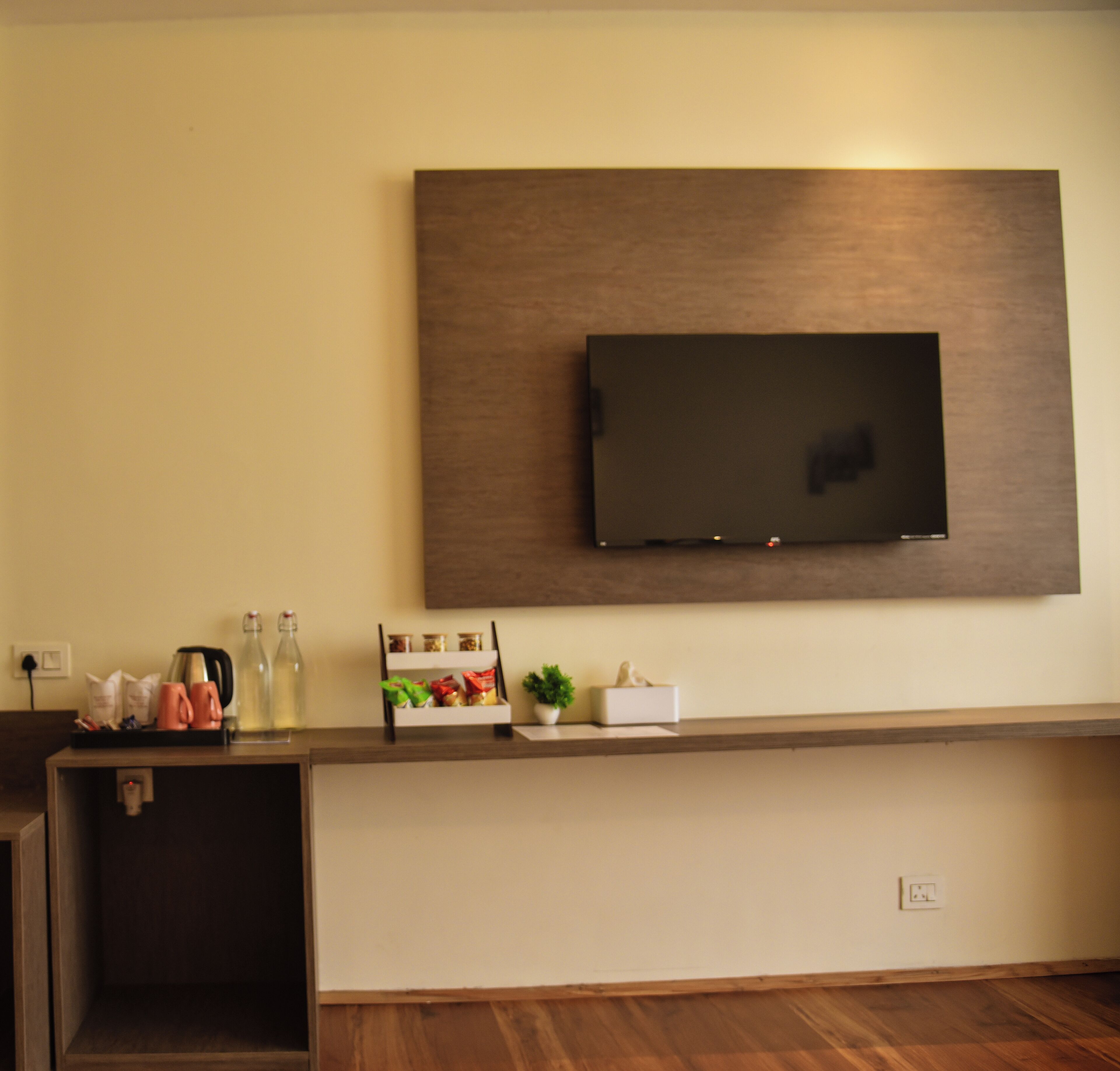 Un área de recepción moderna de hotel con un televisor de pantalla plana montado en la pared, un mostrador de recepción de madera y una planta pequeña sobre el mostrador.