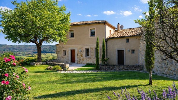 Une maison en pierre à deux étages, avec un toit en tuiles, entourée d’une pelouse bien entretenue et de plantes en fleurs.