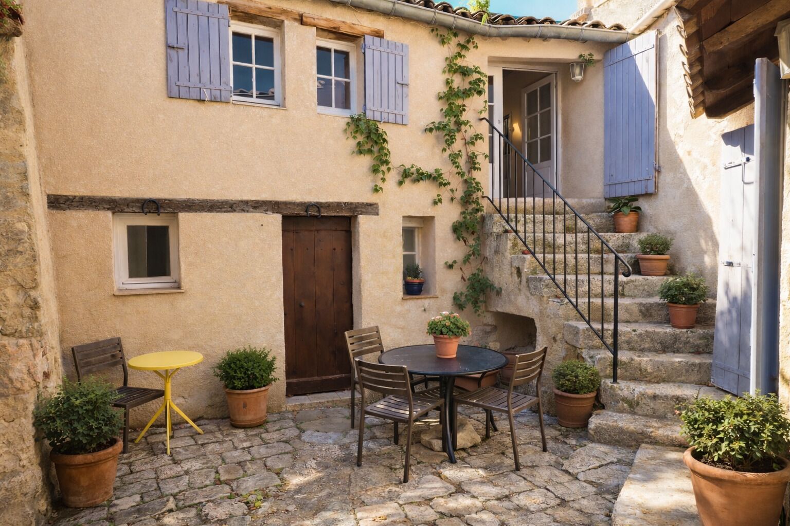 A courtyard with a stone floor, a round table with chairs, potted plants, and a staircase leading to an upper level.