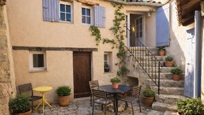 Une cour avec un sol en pierre, une table ronde entourée de chaises, des plantes en pot et un escalier menant à un niveau supérieur.