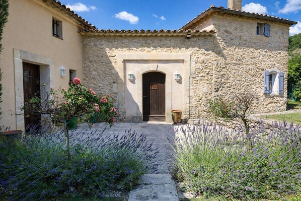 Un edificio de piedra con una entrada central, rodeado de plantas de lavanda y un pequeño árbol.