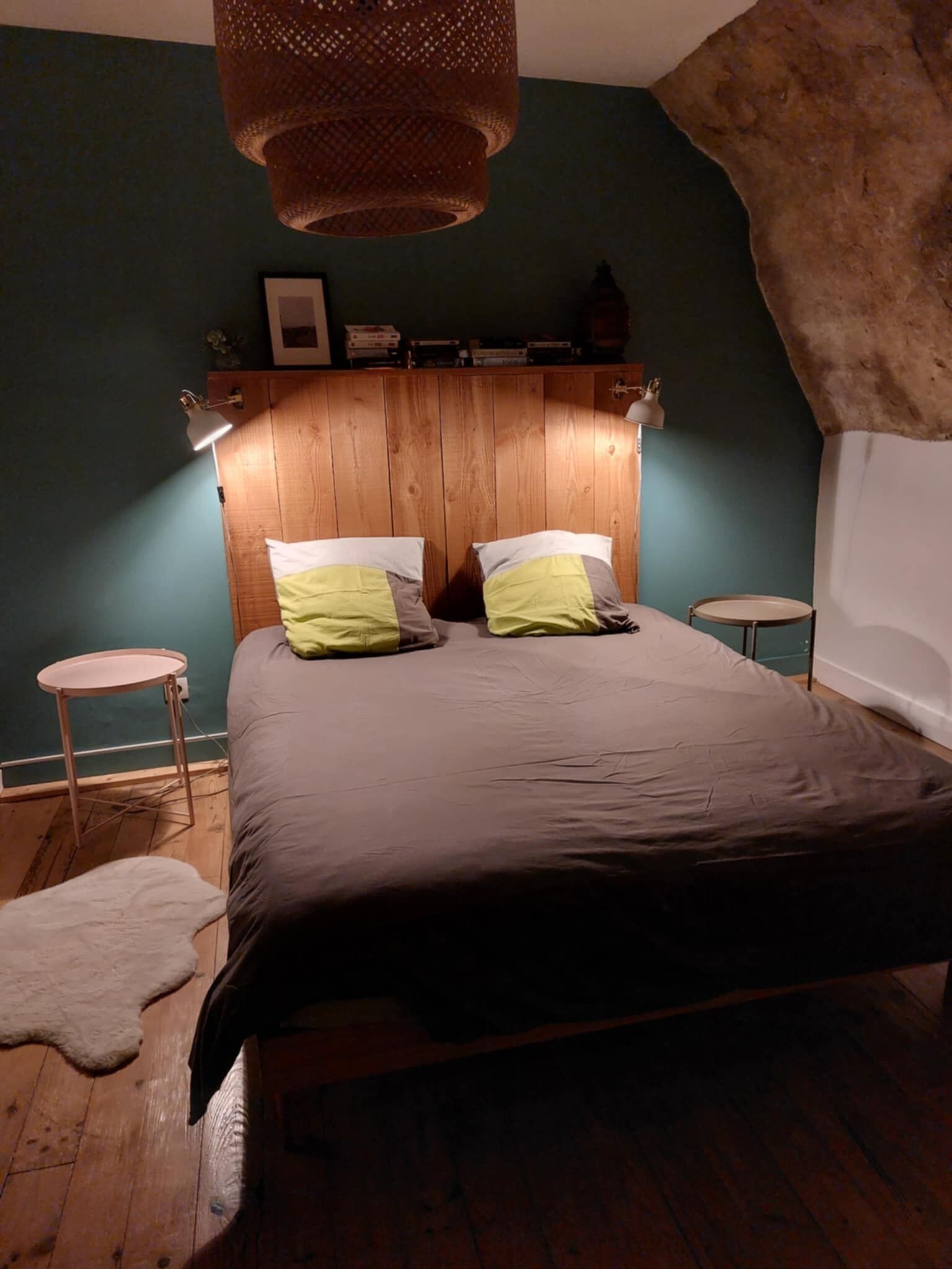A bedroom with a wooden headboard, two bedside tables, a bed with grey and yellow pillows, a hanging light fixture, and a white wall.