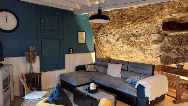 Een gezellige woonkamer met een bank, een salontafel en een gele fauteuil. Er hangt een klok aan de muur en achter de bank bevindt zich een stenen muur.