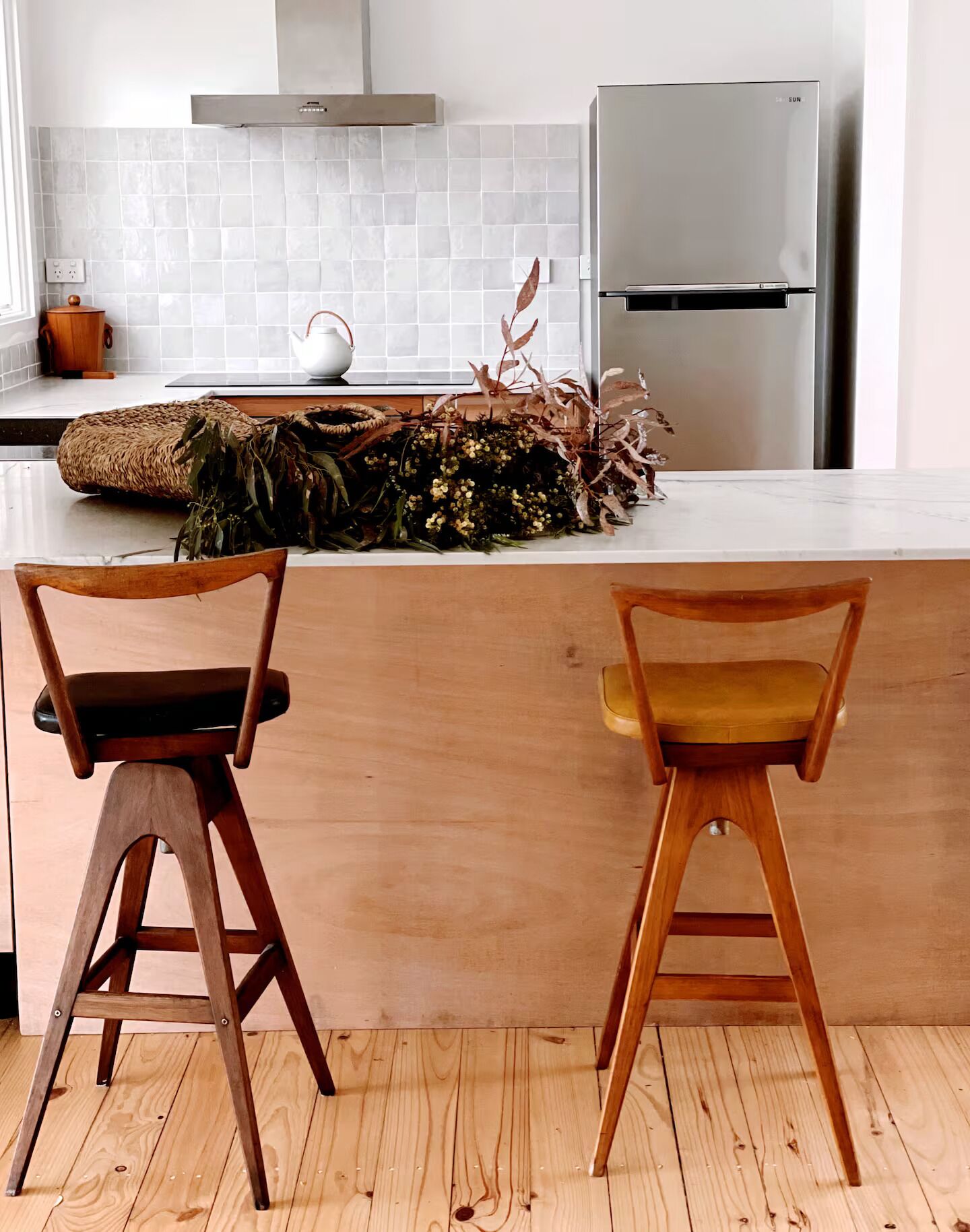 A kitchen with a white tiled backsplash, a stainless steel refrigerator, and two wooden chairs with cushioned seats.
