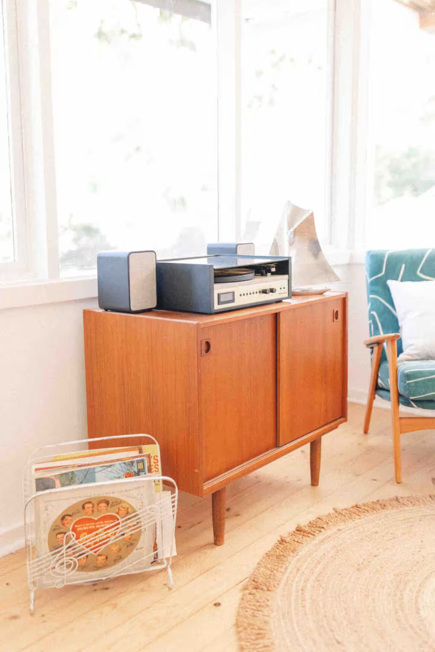 Un meuble en bois contenant une platine vinyle et des haut-parleurs, un porte-revues et un fauteuil de couleur turquoise.