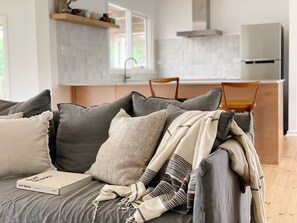En moderne stue med en grå sofa, en bog med titlen 'Surf Shacks', og et køkken i baggrunden.