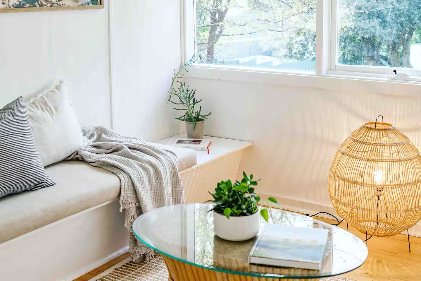 A cozy living room with a sofa, a glass coffee table with a plant, a wicker lamp, and a window with a view of trees.