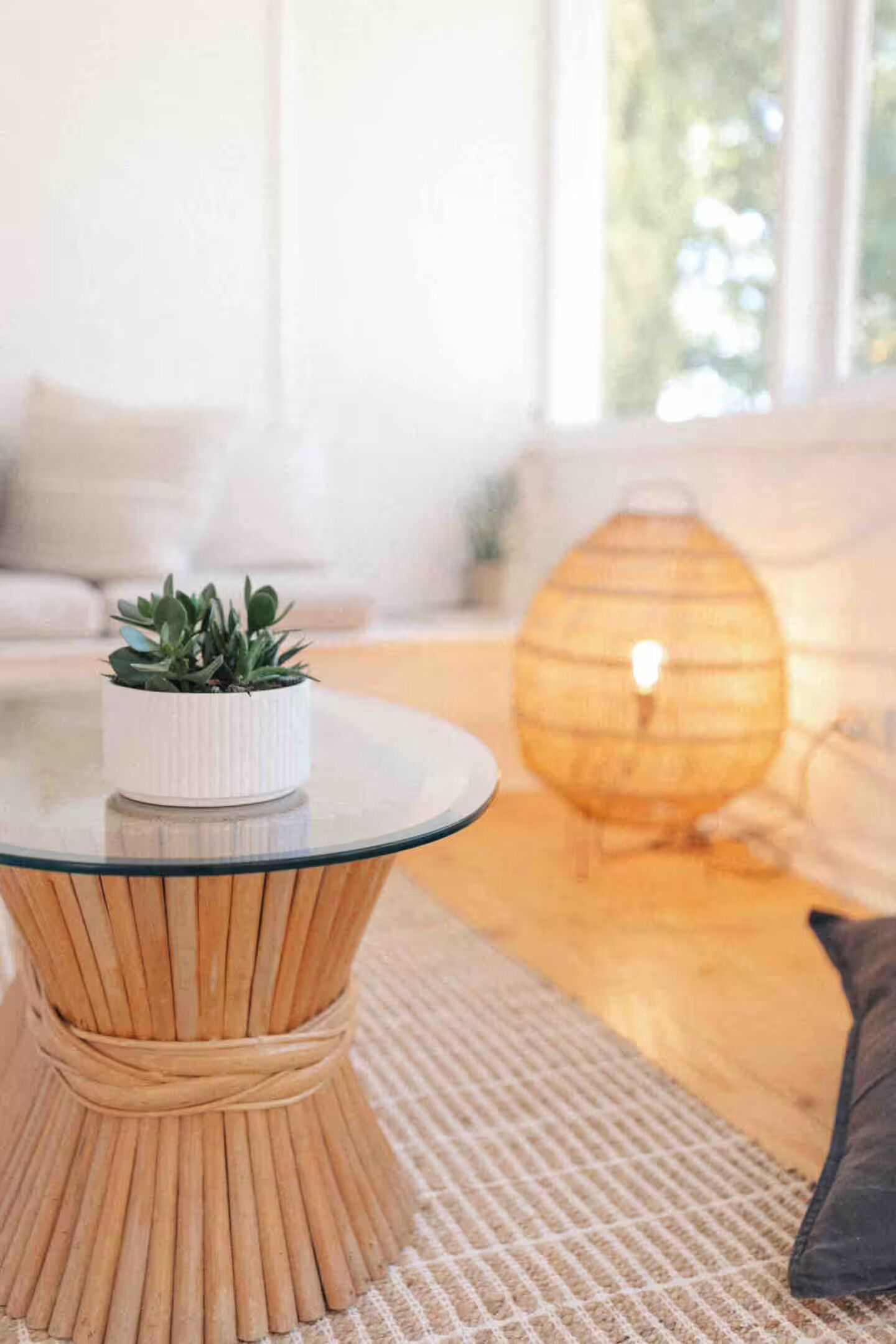 A round glass coffee table with a woven base, a small potted plant, and a woven lamp on a textured rug.