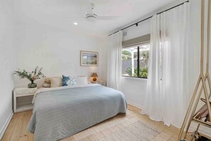 Ein Schlafzimmer mit Bett, Nachttischen, einer Lampe, einem Fenster mit Vorhängen und einem Deckenventilator.