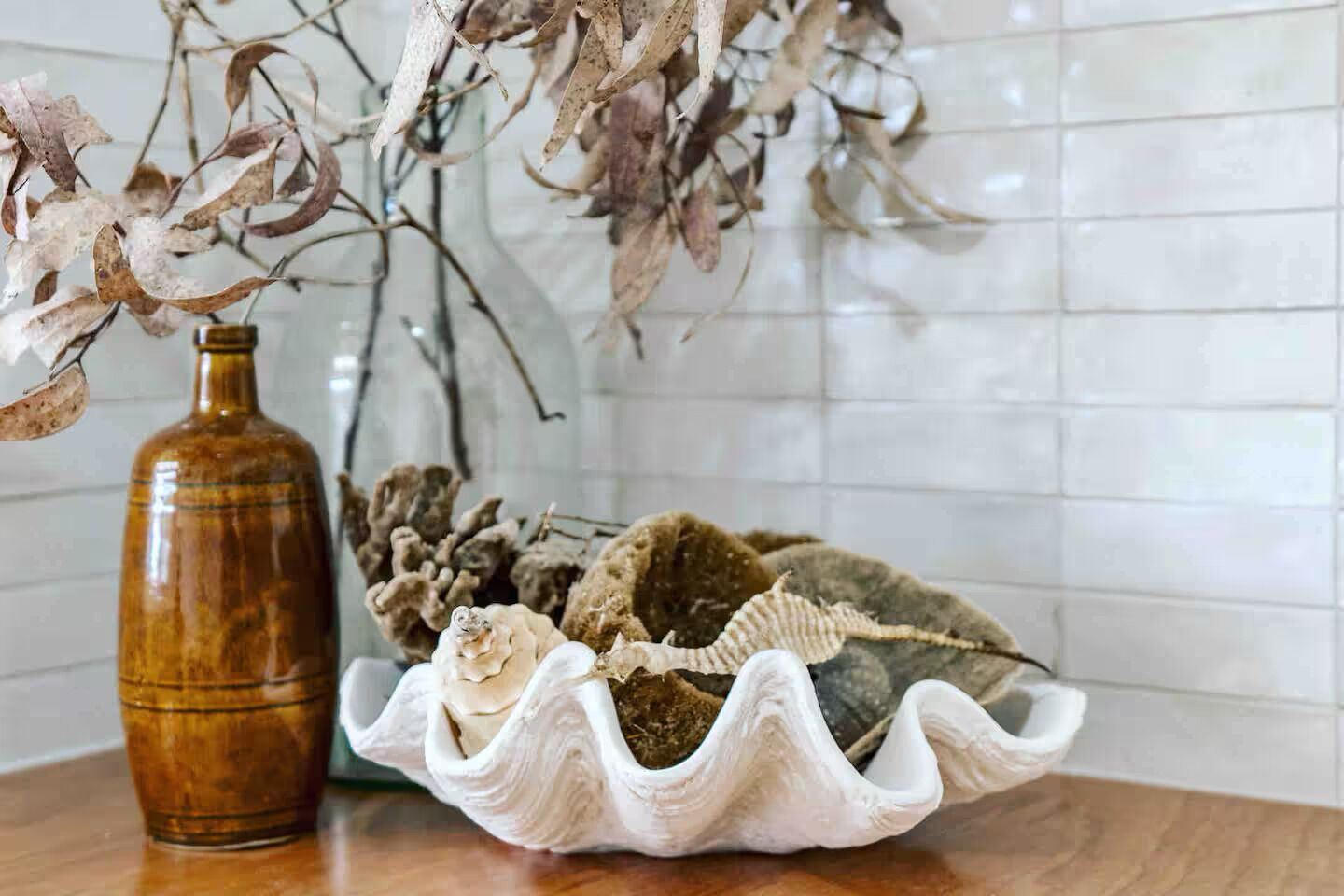 A ceramic vase with dried leaves and a bowl of seashells on a wooden surface.