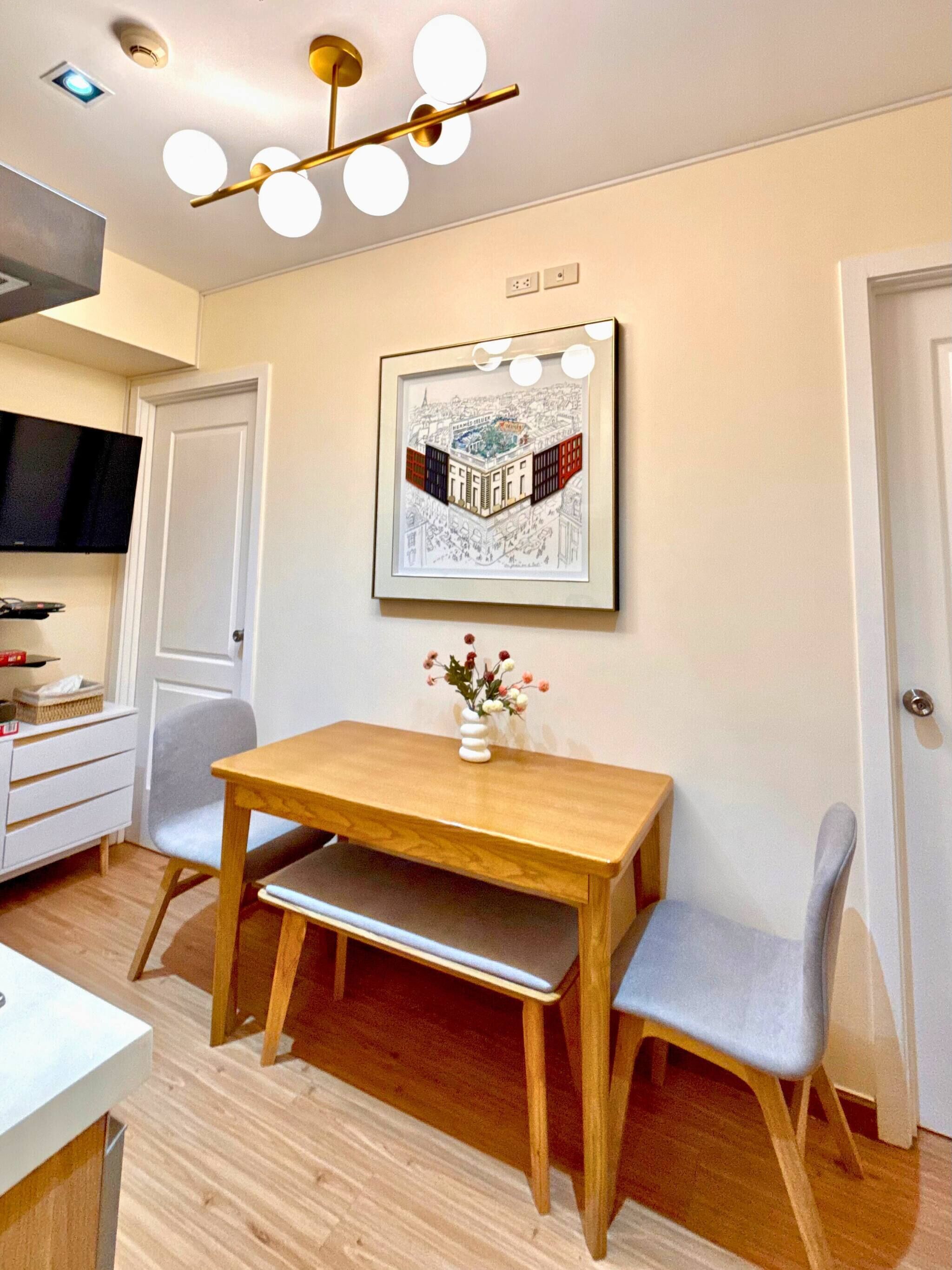 Een eetgedeelte met een houten tafel en twee stoelen, een stadsgezicht in een lijst boven de tafel, en een televisie aan de muur.