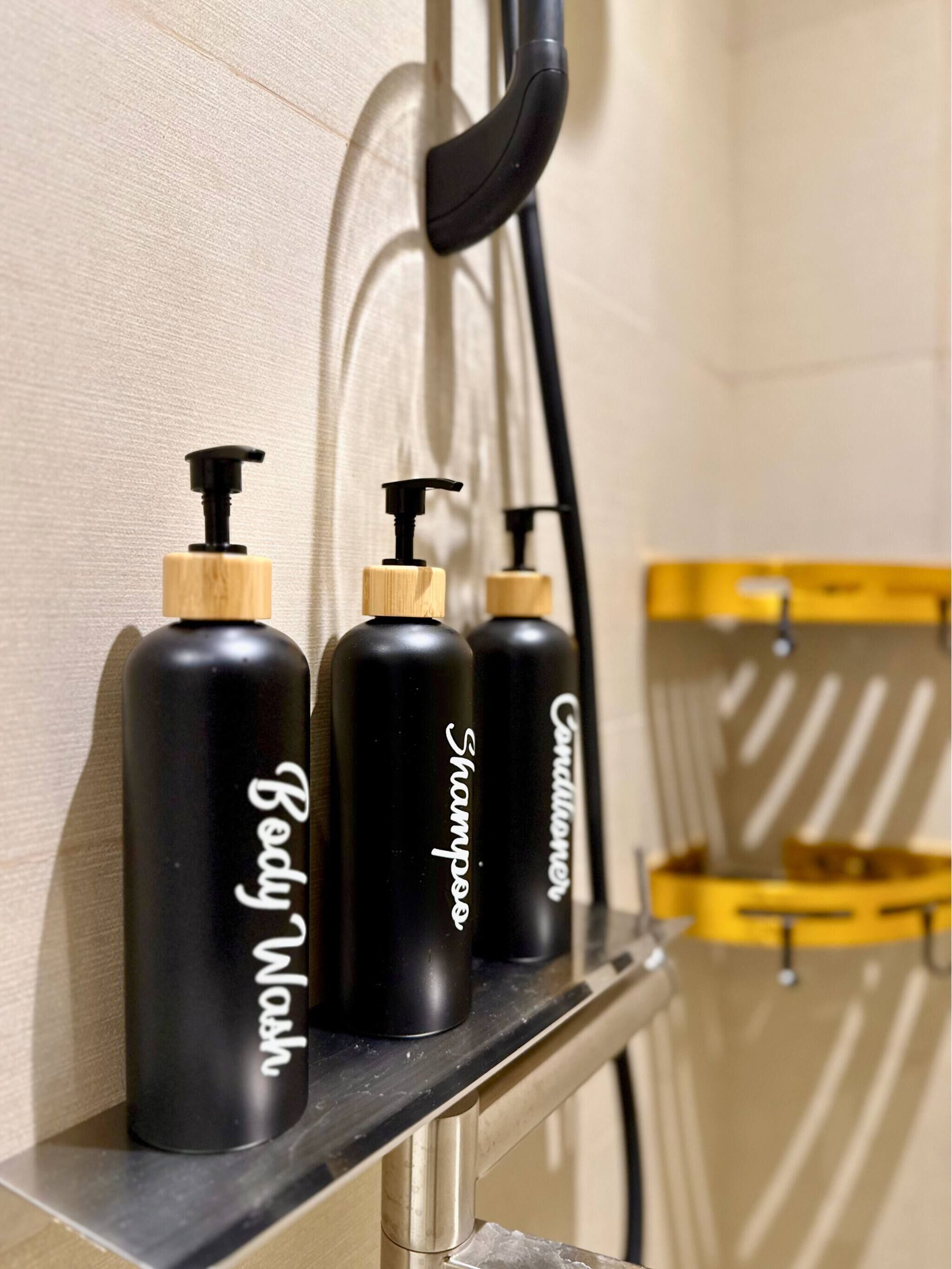 Drei schwarze Flaschen mit Holzkorken, beschriftet mit 'Body Wash', 'Shampoo' und 'Conditioner', auf einem Regal.