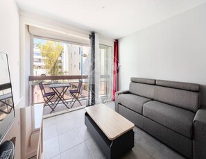 A modern living room with a grey sofa, a wooden coffee table, and a balcony with a table and chairs. - The Commodore Residence (Le Grau-Du-Roi)