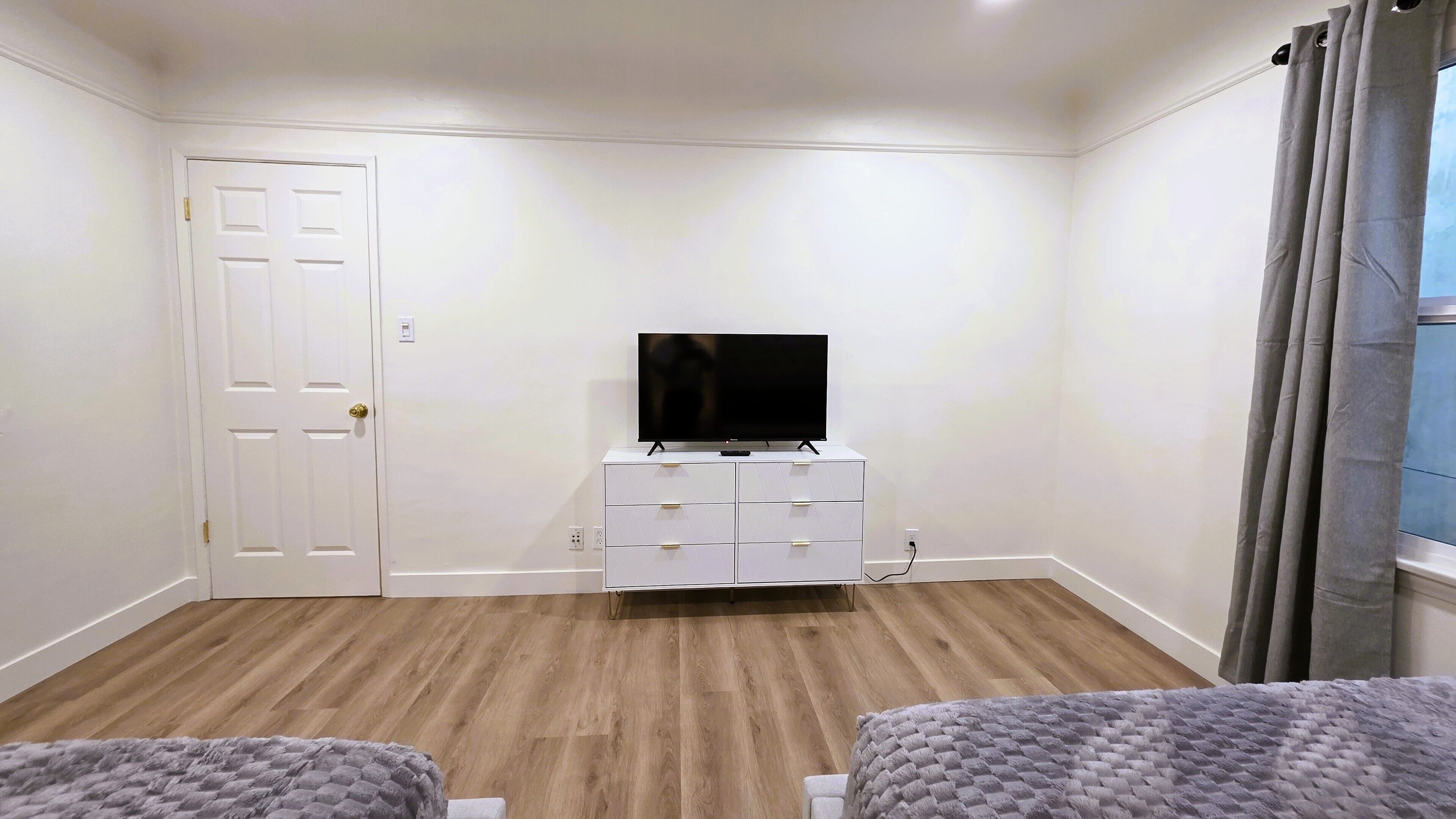 Una habitación con una puerta blanca, un televisor de pantalla plana sobre un aparador blanco, suelo de madera y dos camas grises.