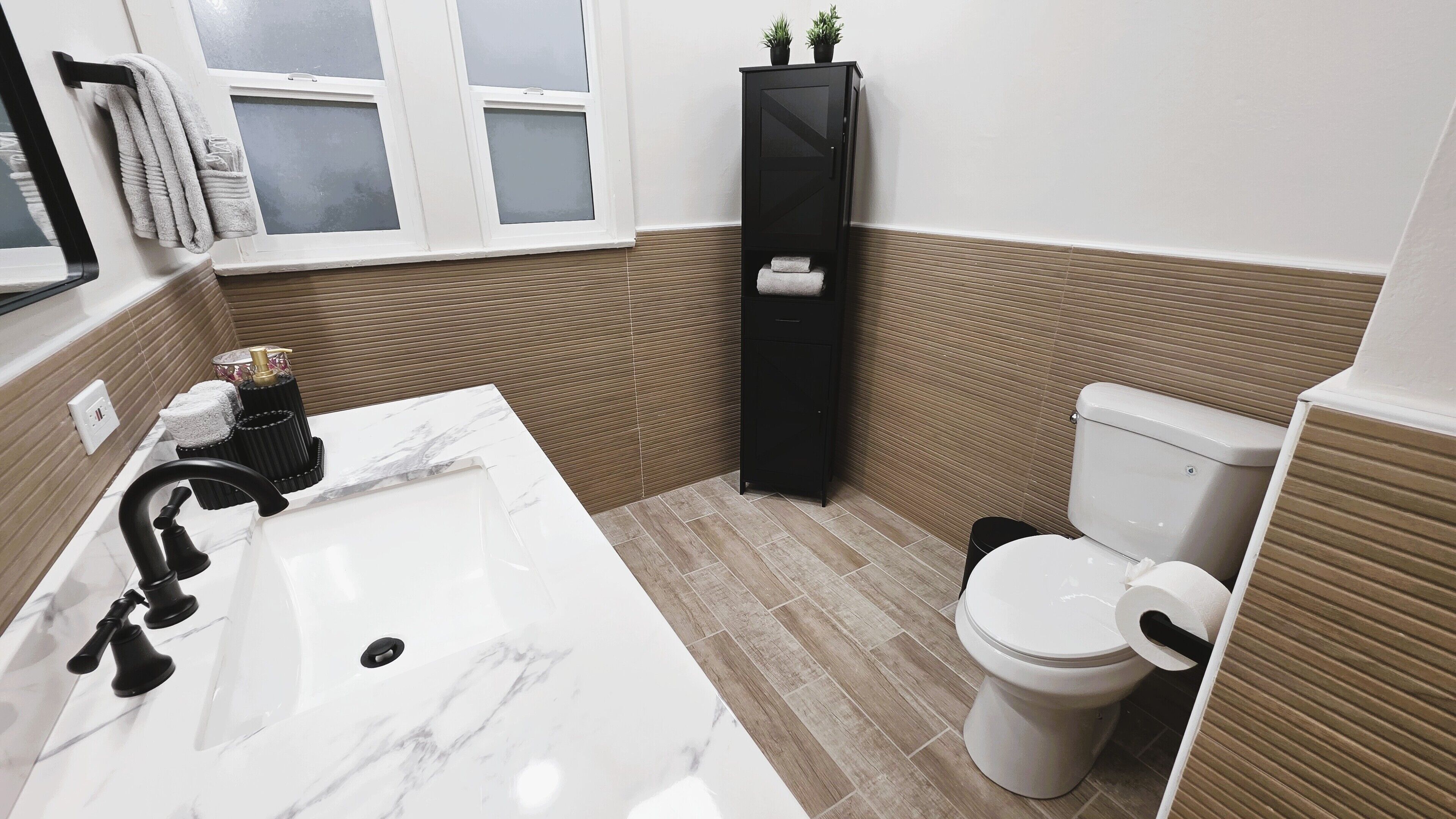 A bathroom with a white marble sink, black faucet, and a toilet. There is a towel rack with towels and a black cabinet with plants on top.
