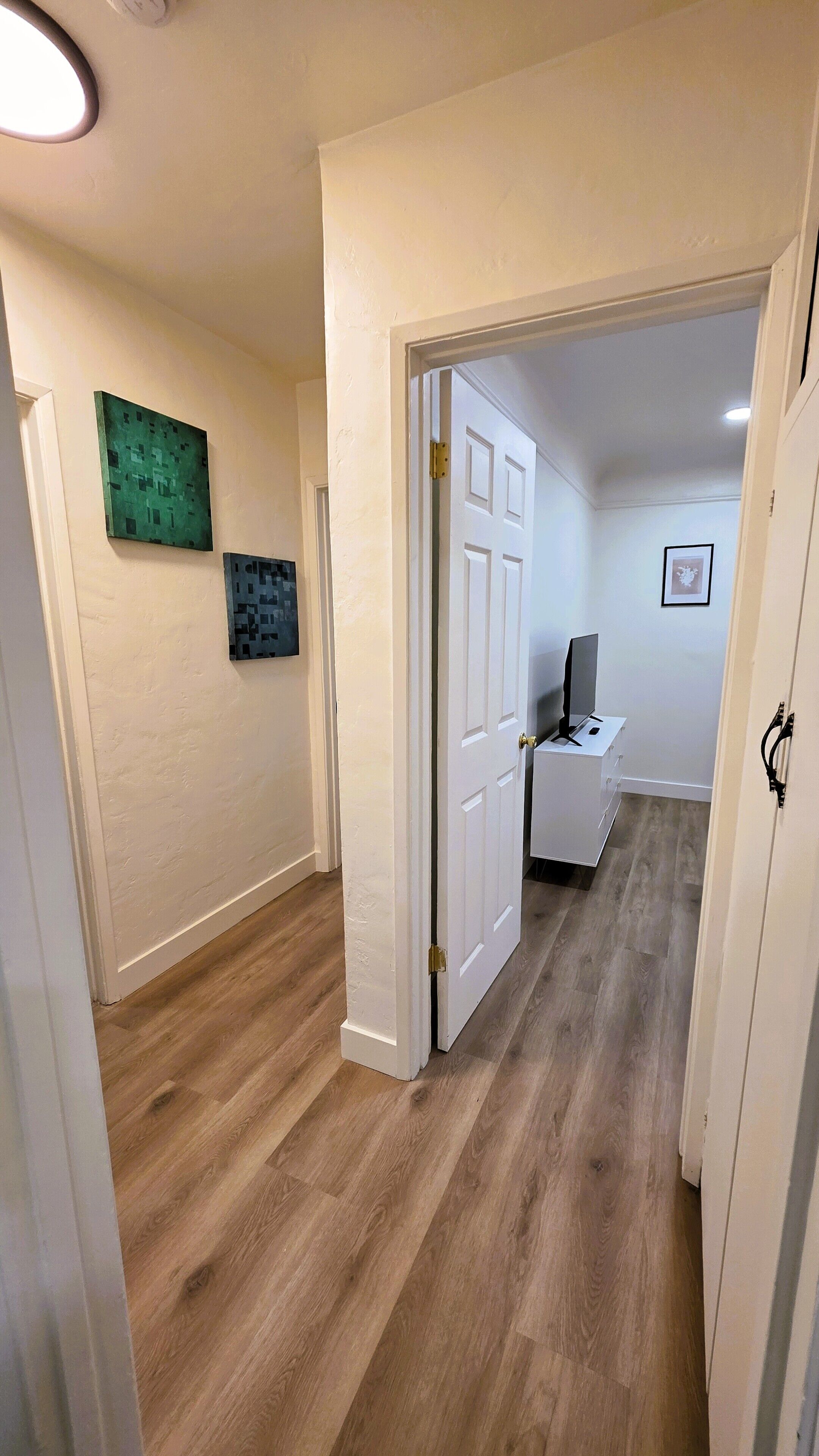 Un couloir doté d’un plancher en bois, de murs blancs et d’une porte donnant sur une pièce où se trouvent un bureau et une chaise.
