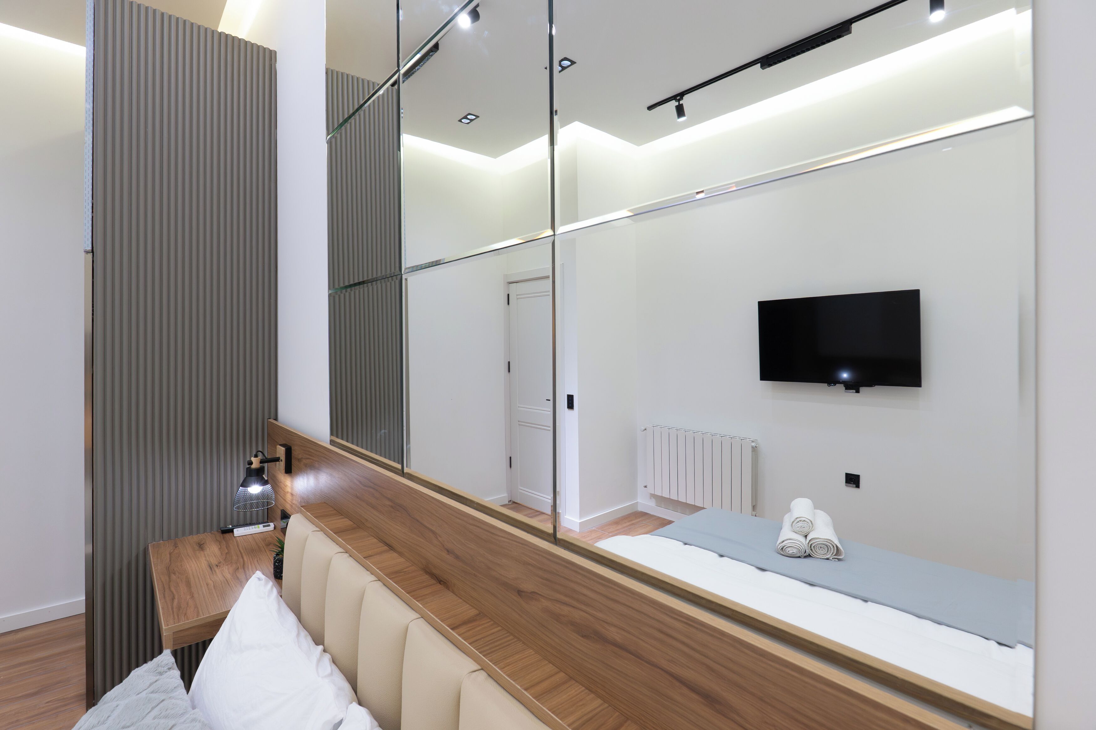 A modern hotel room with a large mirror, a flat-screen TV, a wooden bedside table, and a bed with white bedding.