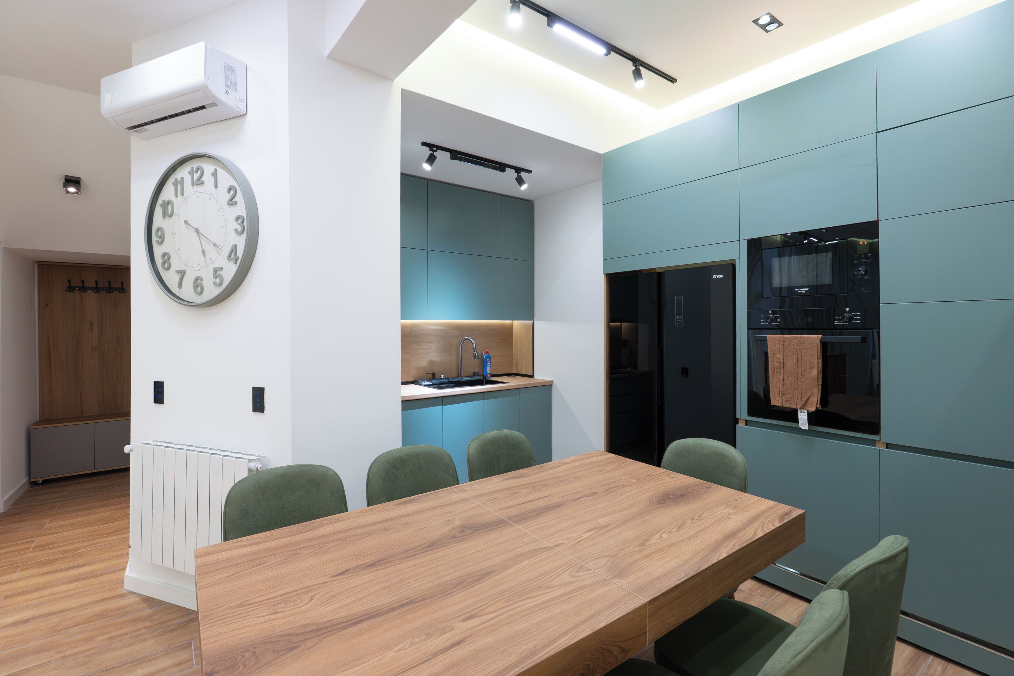 Une cuisine moderne dotée d’un four encastré, d’une horloge au mur et d’un coin repas avec une table en bois et des chaises vertes.
