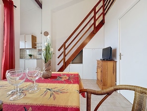 A dining area with a table set for two, a wooden chair, a small cabinet with a television, and a staircase leading to an upper floor. - Le Millmer by Interhome (Saint Aygulf)