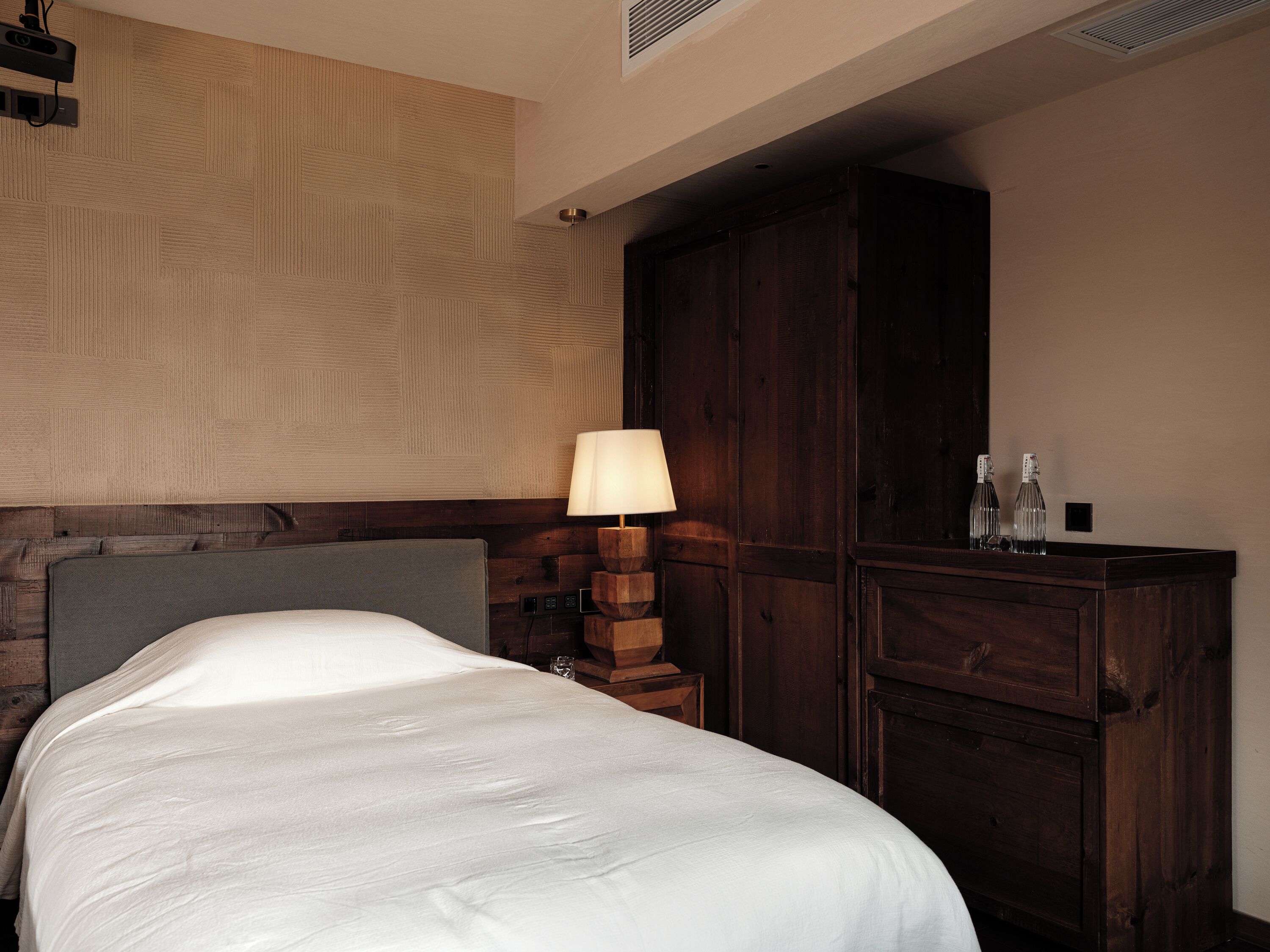 Una habitación de hotel con una cama, cabecera de madera, mesita de noche con lámpara y armario de madera.