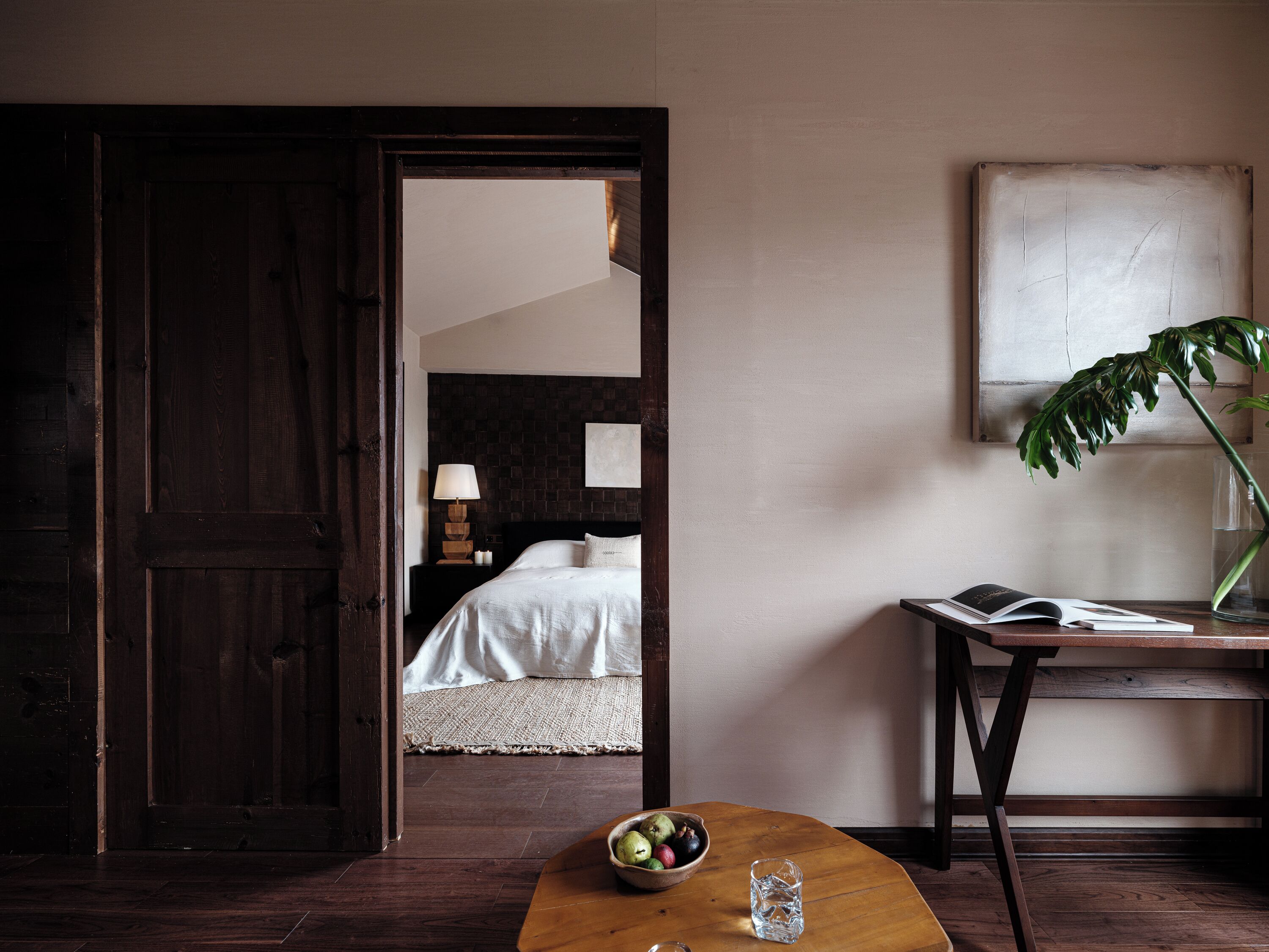 A room with a bed, a nightstand, a table with a bowl of fruit, a glass of water, and a potted plant.