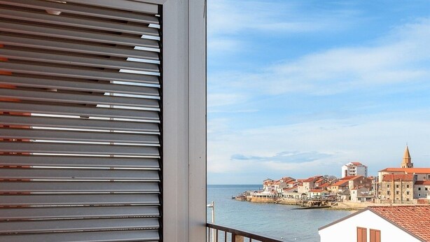 A balcony with horizontal blinds overlooking a coastal town with buildings and a church. - Ares 14 - Modern apartment with sea view, quiet location and close to the beach 20 m. (Umag)