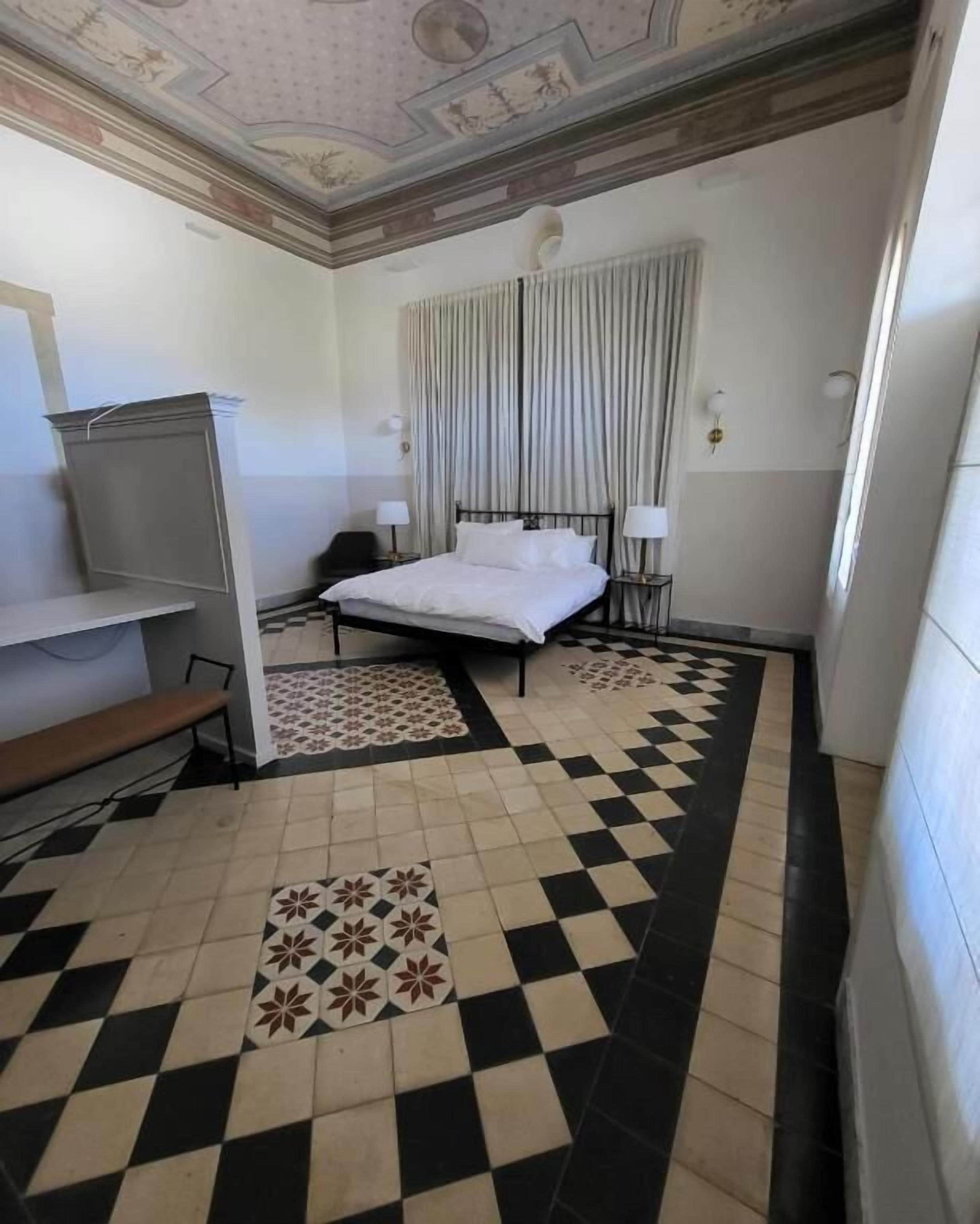A hotel room with a bed, a bench, a desk, and a ceiling with decorative artwork.