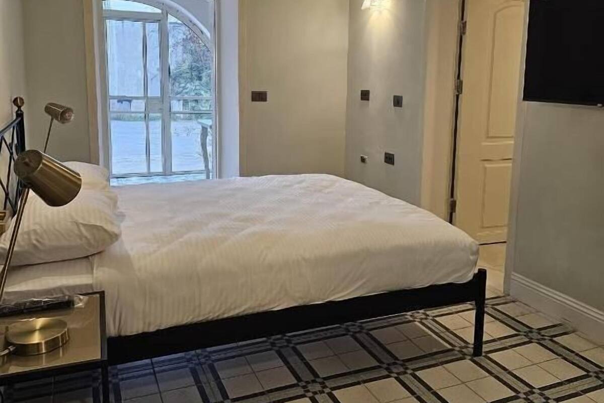 Un dormitorio con una cama, una lámpara de noche, una ventana, una puerta y un televisor montado en la pared.