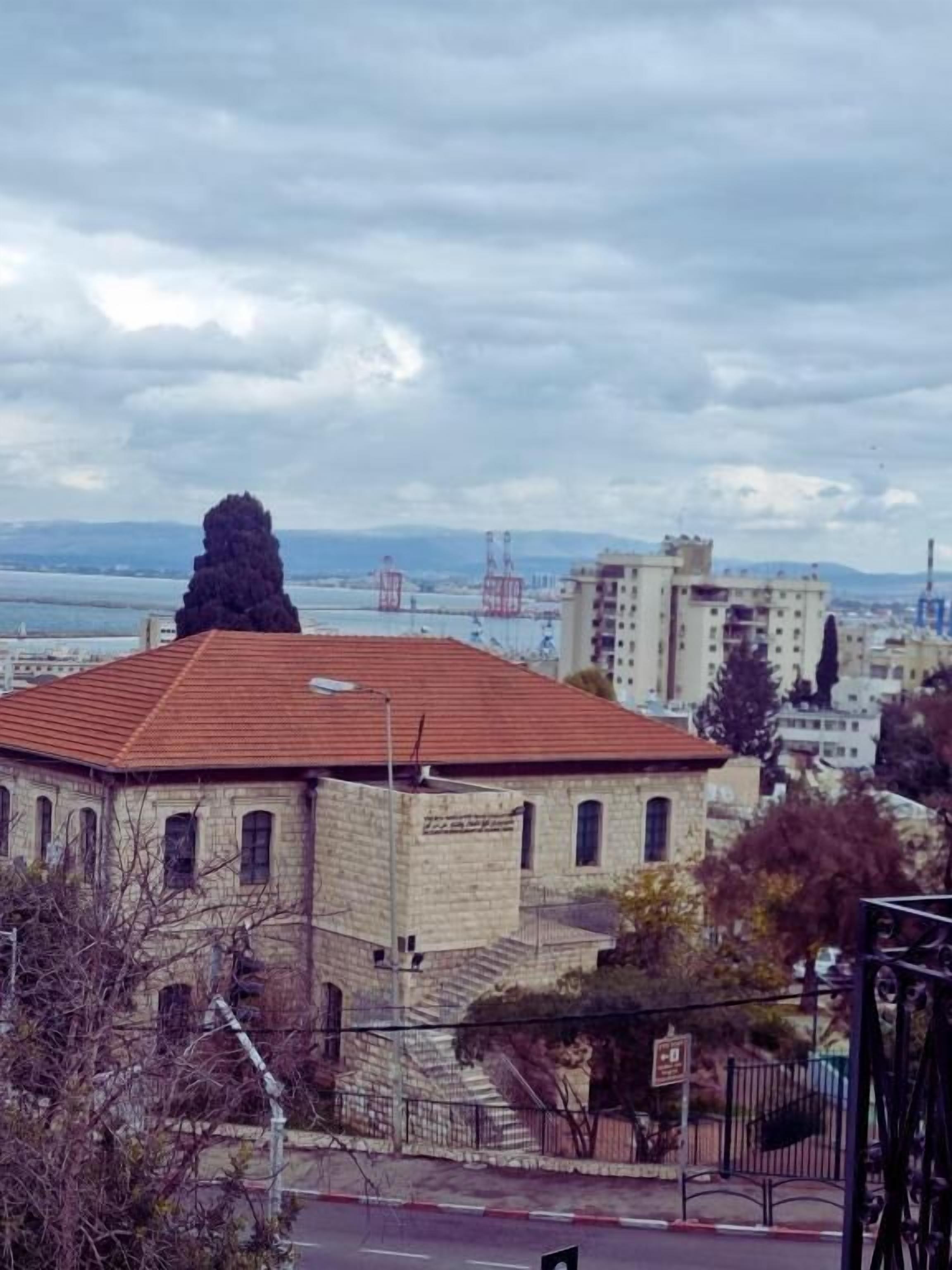 A building with a red-tiled roof and stone walls, located near a body of water with industrial structures in the background.
