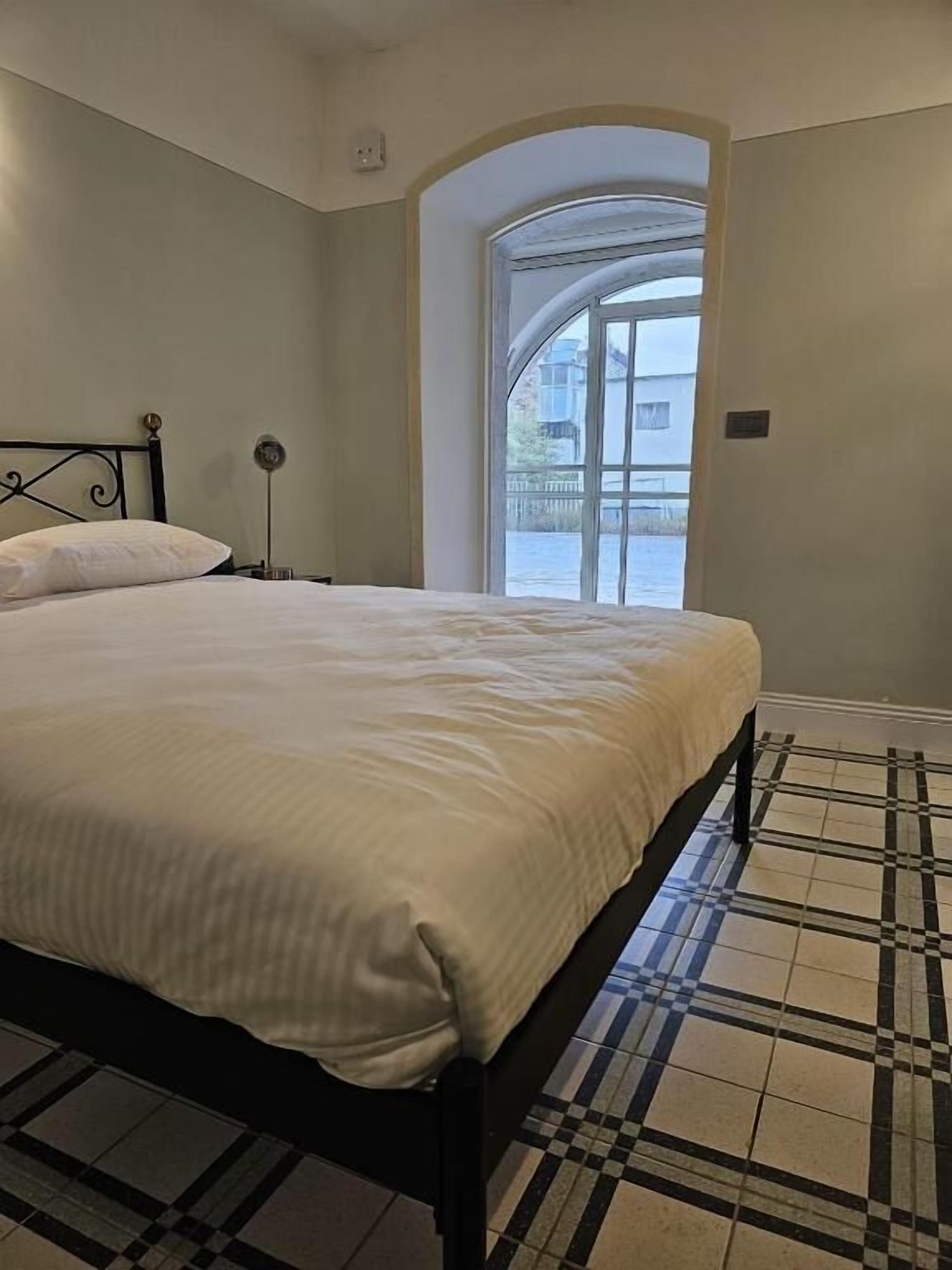 A bedroom with a bed, a nightstand, a lamp, and a view of the outside through a window.