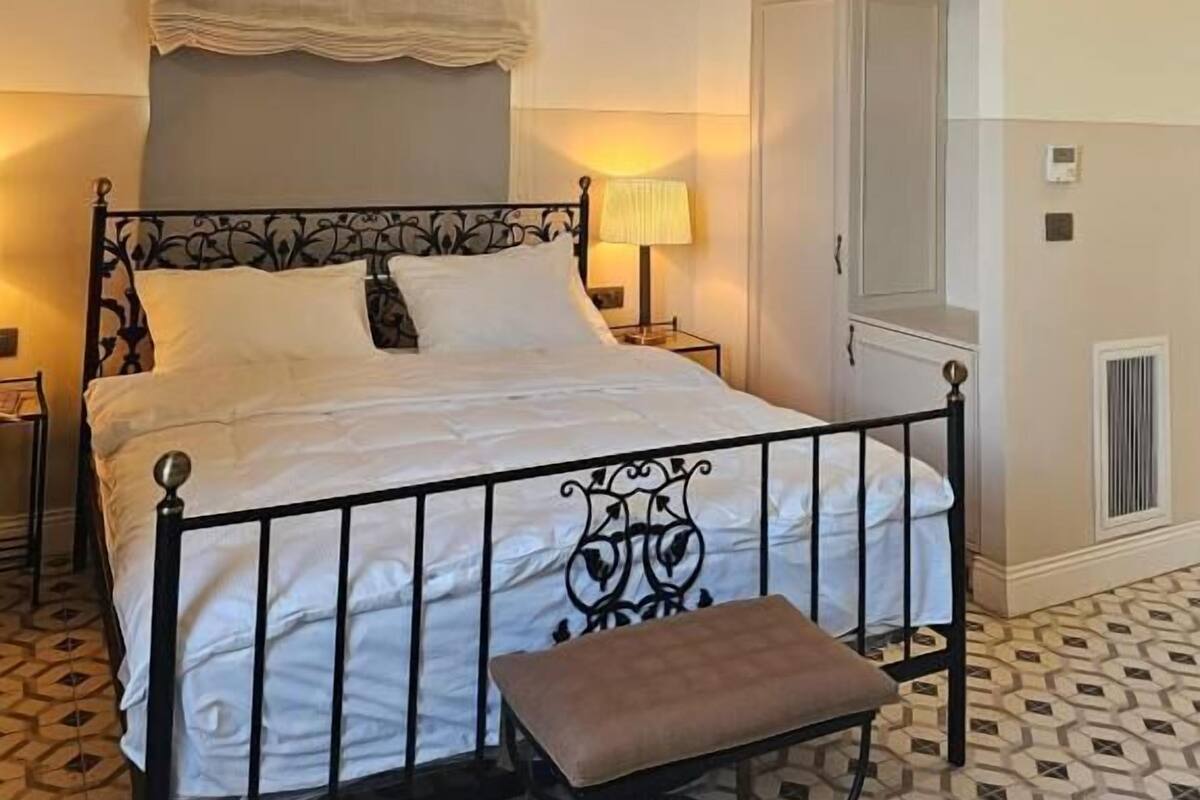 Una cama bien hecha con un cabecero, una lámpara de noche y un banco al pie de la cama.
