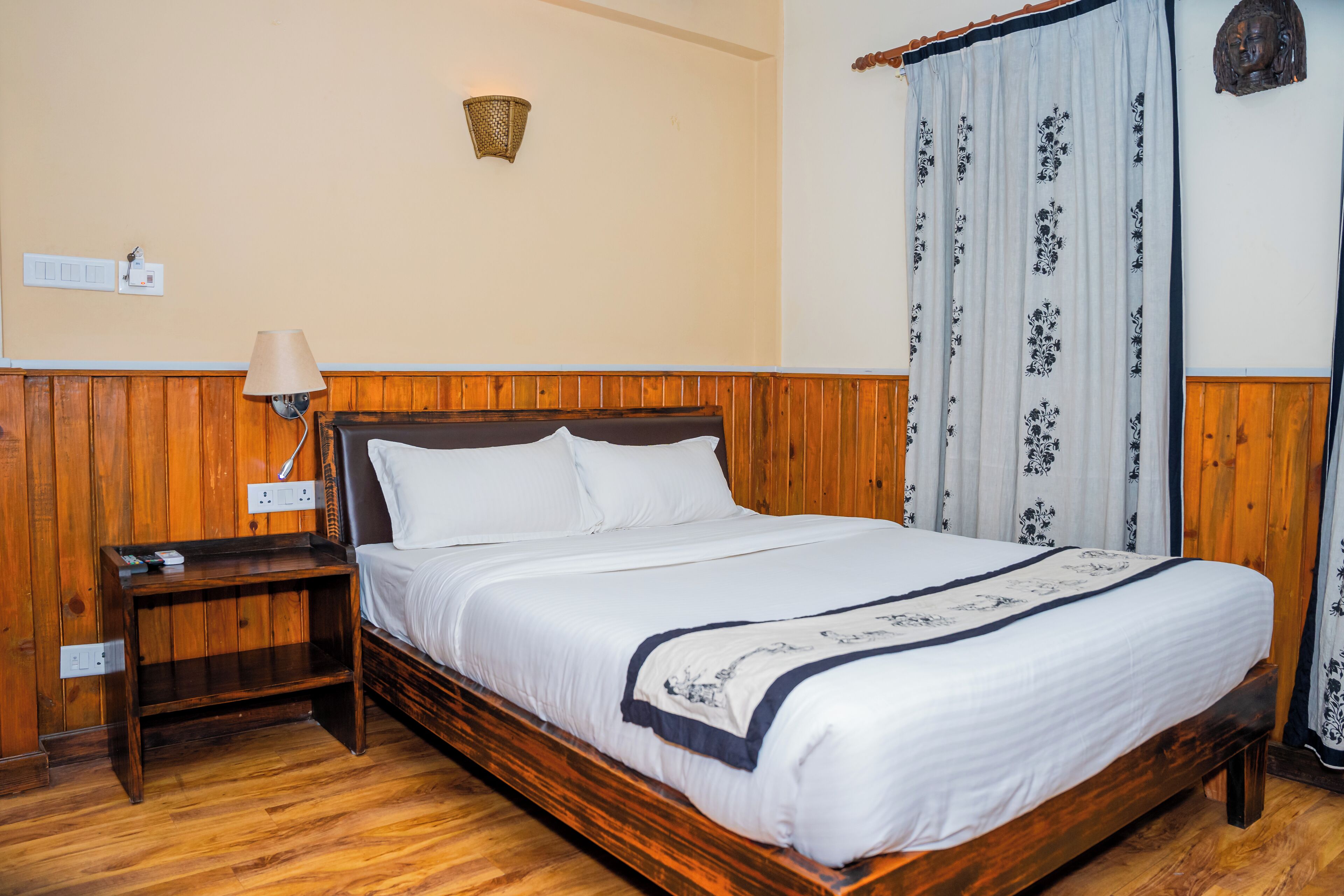 A wooden-framed bed with white bedding, a bedside table, a lamp, and a wall-mounted light fixture.