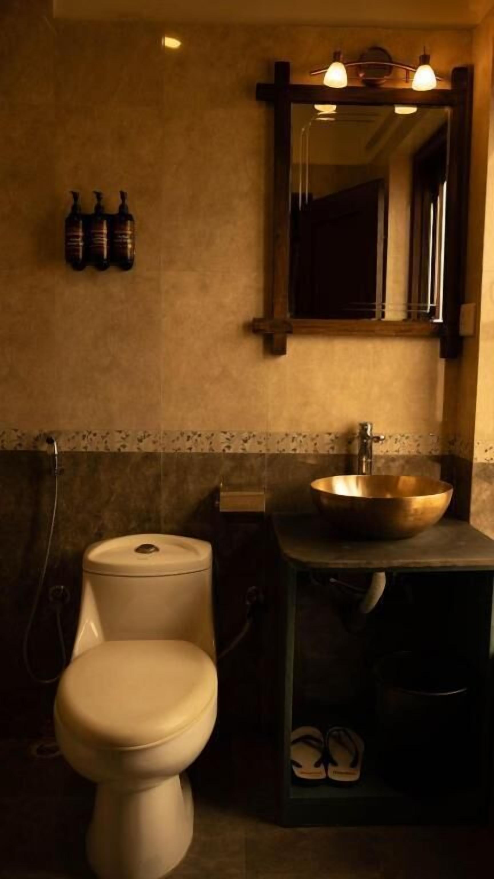 Un baño con inodoro, lavabo y espejo sobre él. Hay botellas en la pared y un par de pantuflas debajo del lavabo.