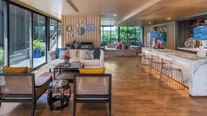 A modern living room with a sofa, coffee table, and chairs, a kitchen area with a bar, and large windows offering a view of greenery. - Friendly 2BR in Phuket Central With Pool & gym (Wichit)