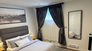 Een slaapkamer met een bed, nachtkastjes, een bedlampje, een groot raam met gordijnen, een spiegel aan de muur en een schilderij.