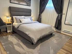 Une chambre moderne avec un lit recouvert d’un tissu gris, une table de chevet avec une lampe, un tapis gris et une grande fenêtre avec des rideaux.
