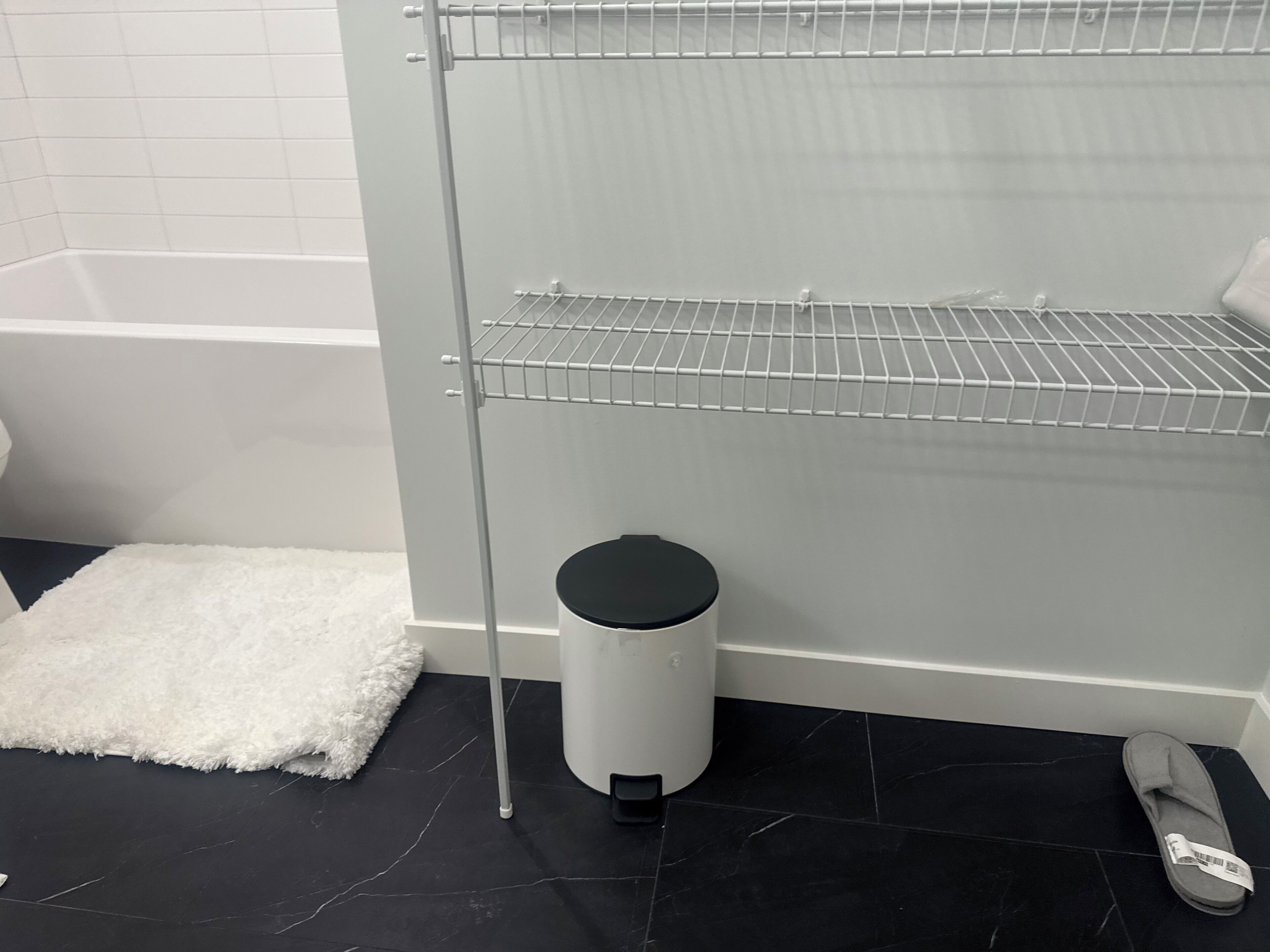 A bathroom with a black tiled floor, a white fluffy rug, a white trash bin, and a pair of slippers.