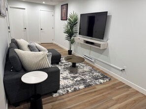 A modern living room with a grey sofa, a small round table, a TV on a wall-mounted stand, and a potted plant. - Fivestar Inspired Private Suite- Downtown Access (Calgary)