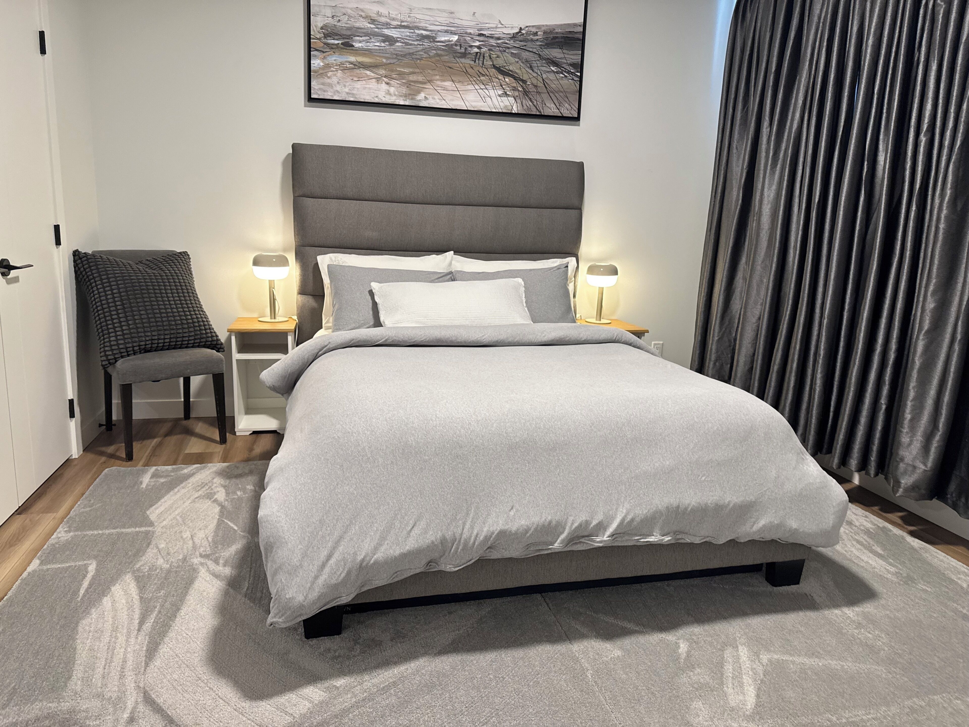 A neatly arranged bedroom with a bed, two bedside lamps, a chair, and a framed picture on the wall.