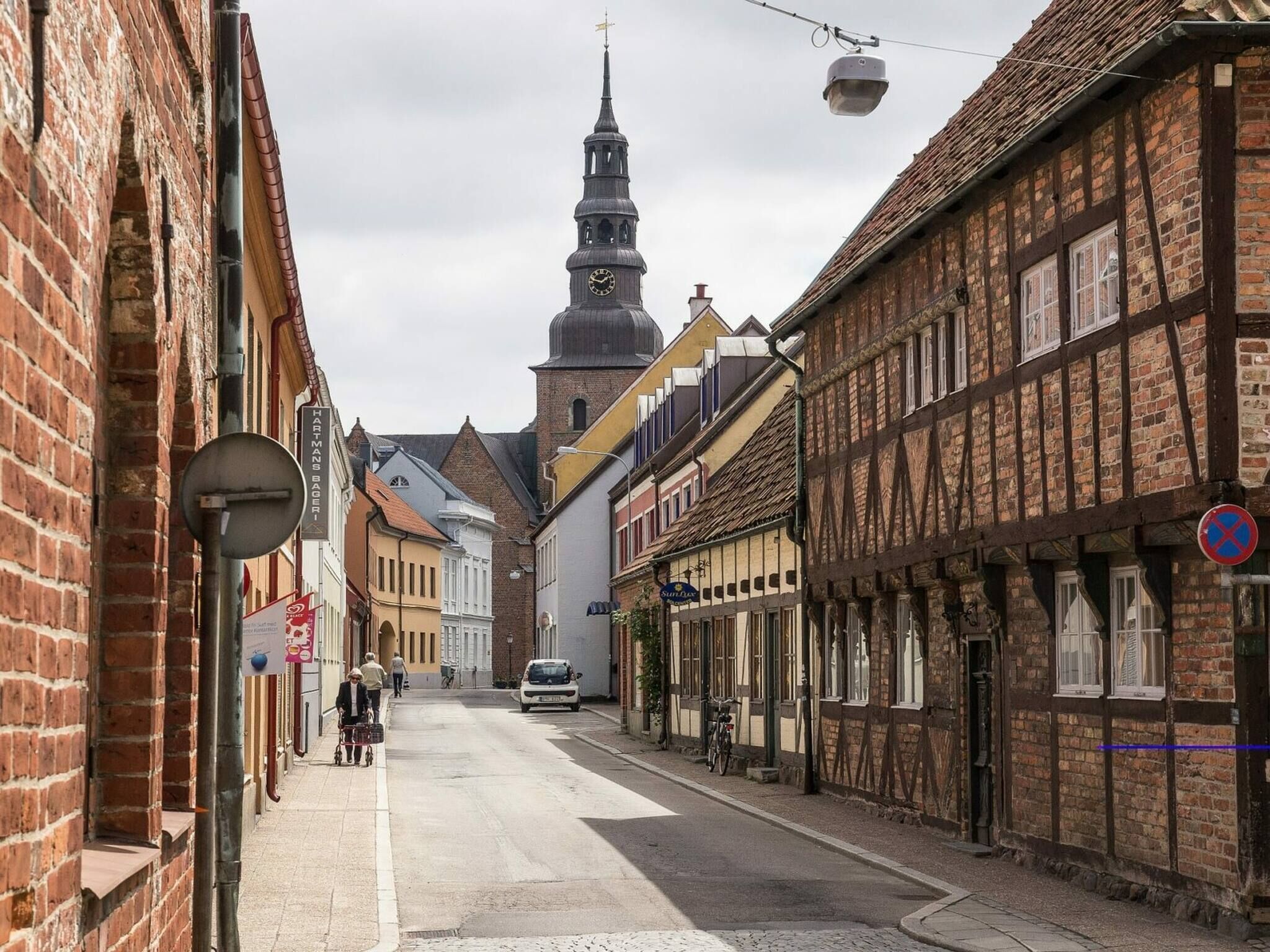 A narrow street lined with traditional half-timbered buildings, a church steeple, and a parked car.