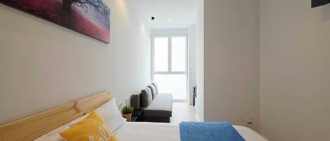 Una habitación de hotel moderna con cama, sofá, un cuadro y un ventanal grande.