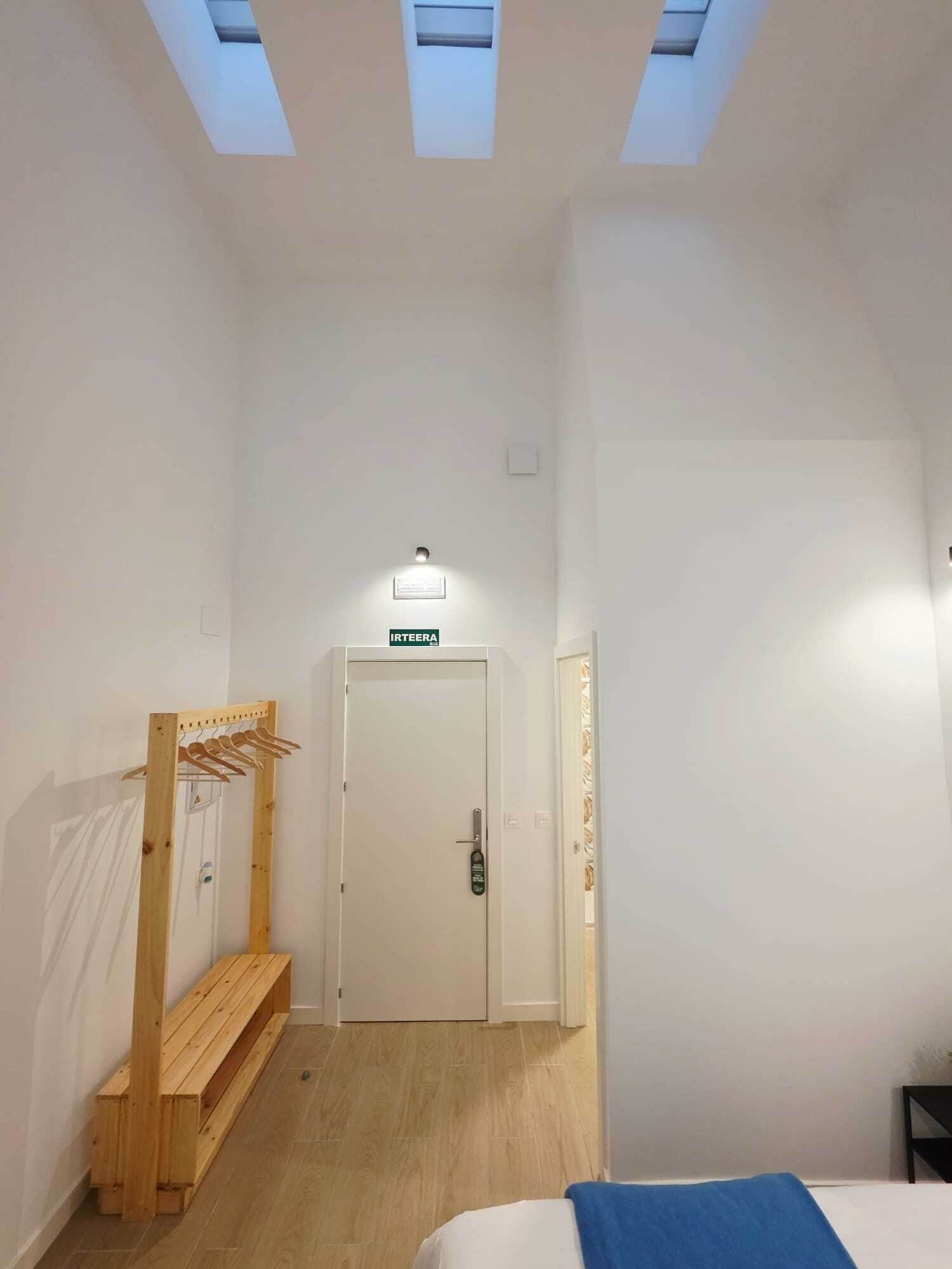 Habitación de hotel con cama, banca, perchero, puerta con cartel y tragaluces.
