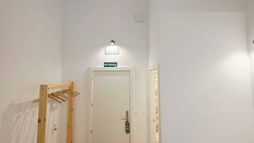 Habitación de hotel con cama, banca, perchero, puerta con cartel y tragaluces.