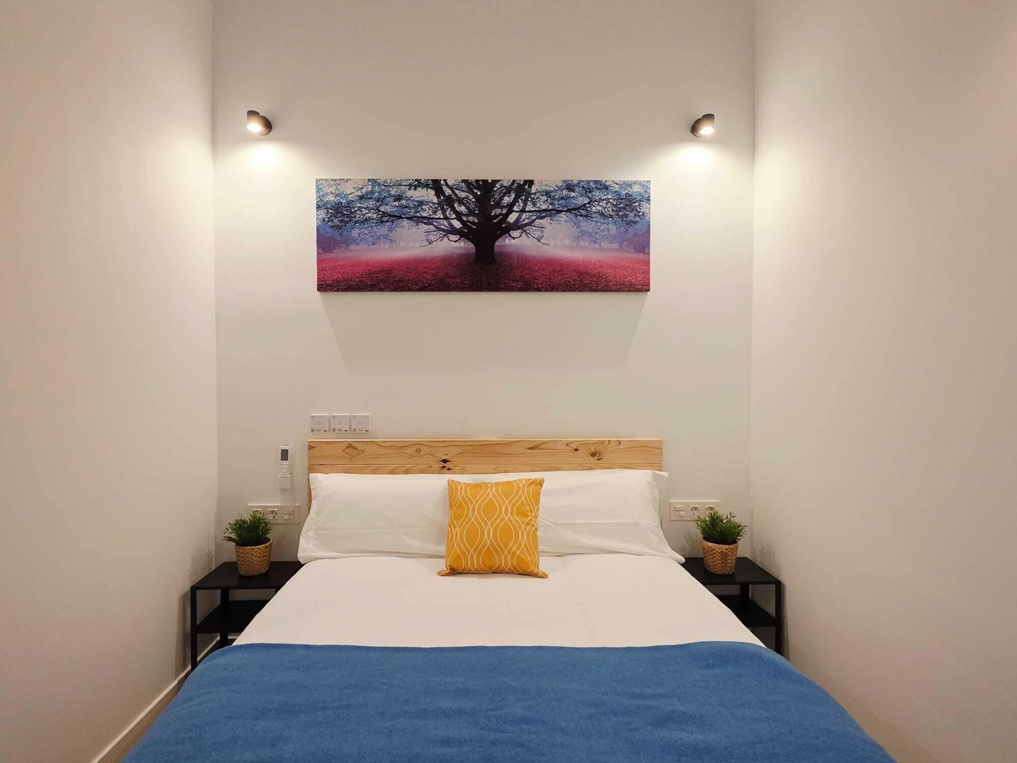 Un dormitorio con cama, un cuadro colgado en la pared, mesitas de noche con plantas y una colcha azul.