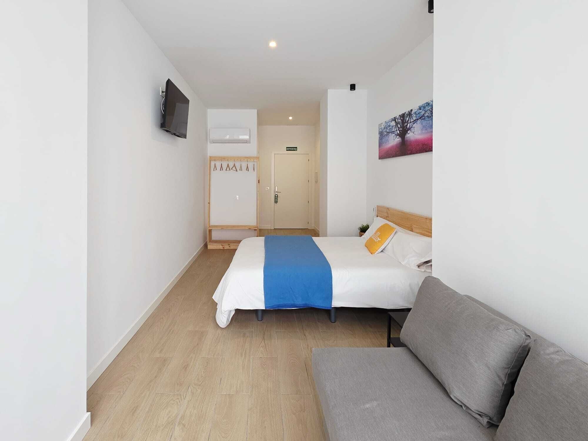A hotel room with a bed, a grey sofa, a TV, a wardrobe, and a painting on the wall.