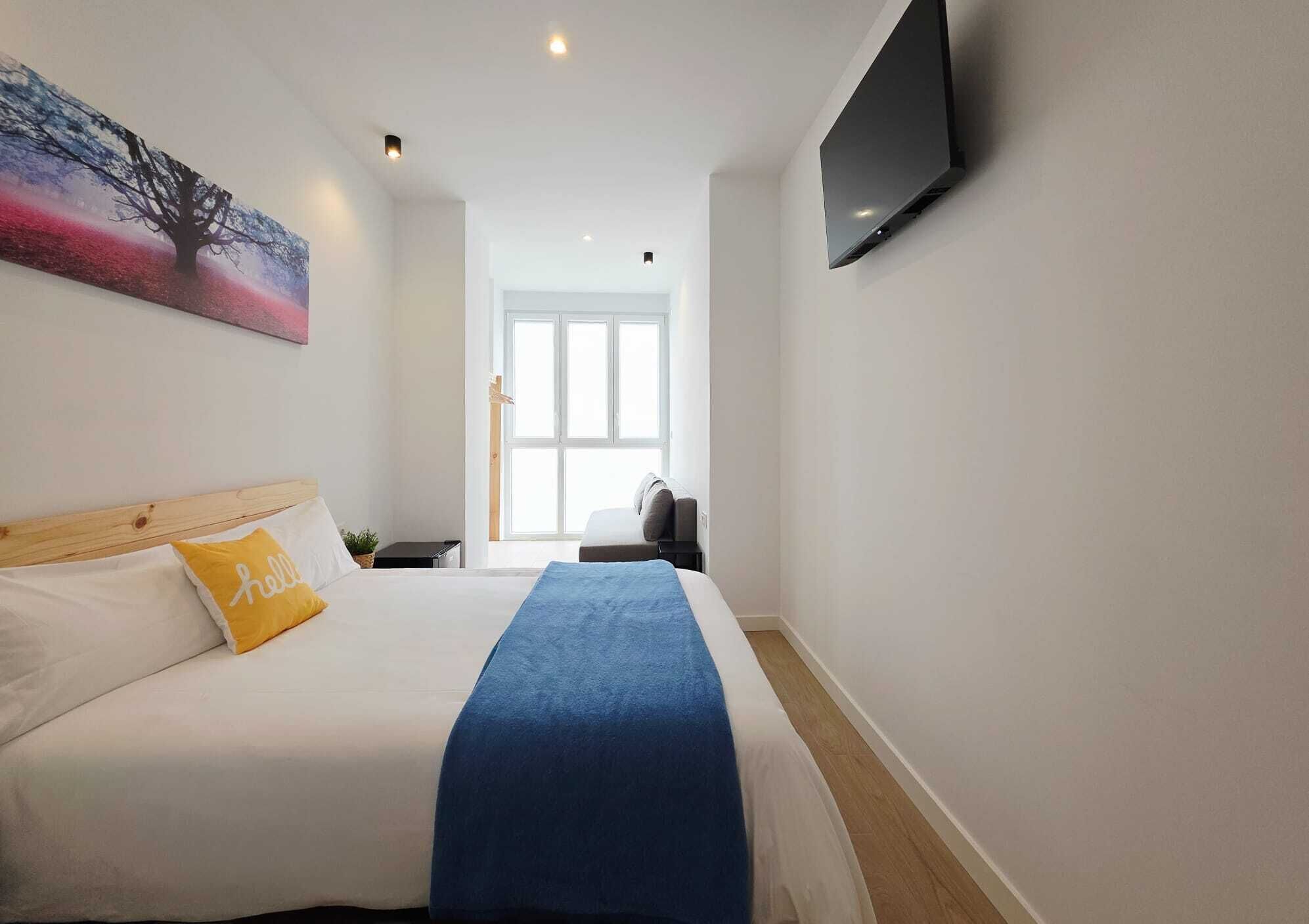 Quarto de hotel com cama, televisão fixada na parede, janela e um quadro sobre a cama.
