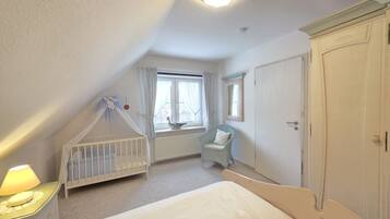 Ein Schlafzimmer mit geneigter Decke, einem Kinderbett, einem Sessel, einem Bett, einem Fenster mit Vorhängen und einem Kleiderschrank.