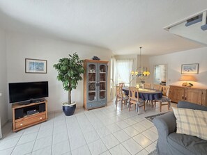 Ein Wohnzimmer mit Flachbildfernseher, Zimmerpflanze, Essbereich mit Tisch und Stühlen sowie einer Couch.